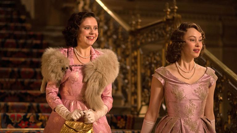 Die beiden Prinzessinnen Margaret (Bel Powley) und Elizabeth (Sarah Gadon) kommen nebeneinander eine prunkvolle Treppe herunter. Sie schauen sich lächelnd um. Beide tragen fast identische, elegante Kleider.