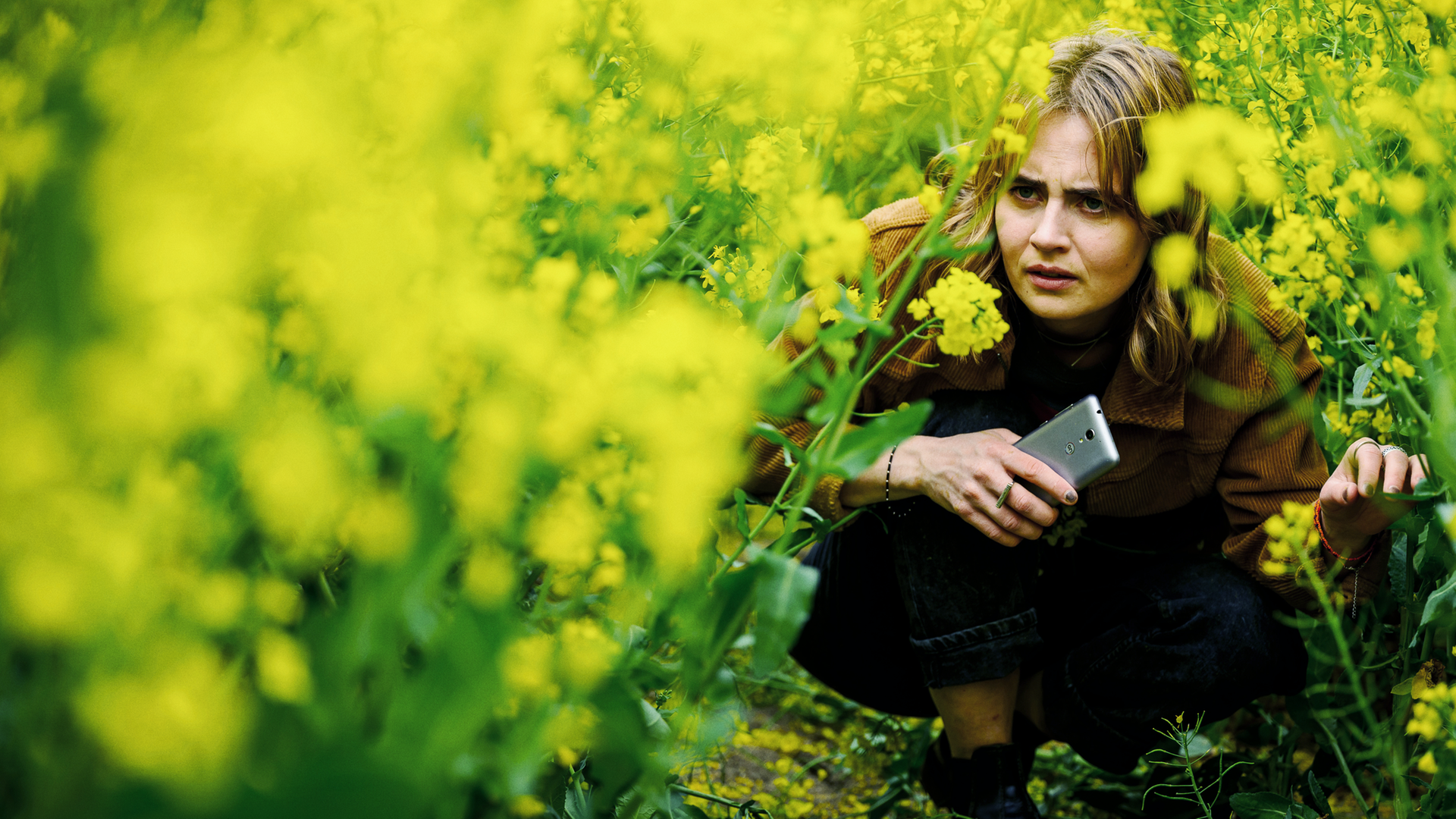 Eine Frau mit einem Handy in der Hand kauert in einem gelb-blühenden Rapsfeld. 