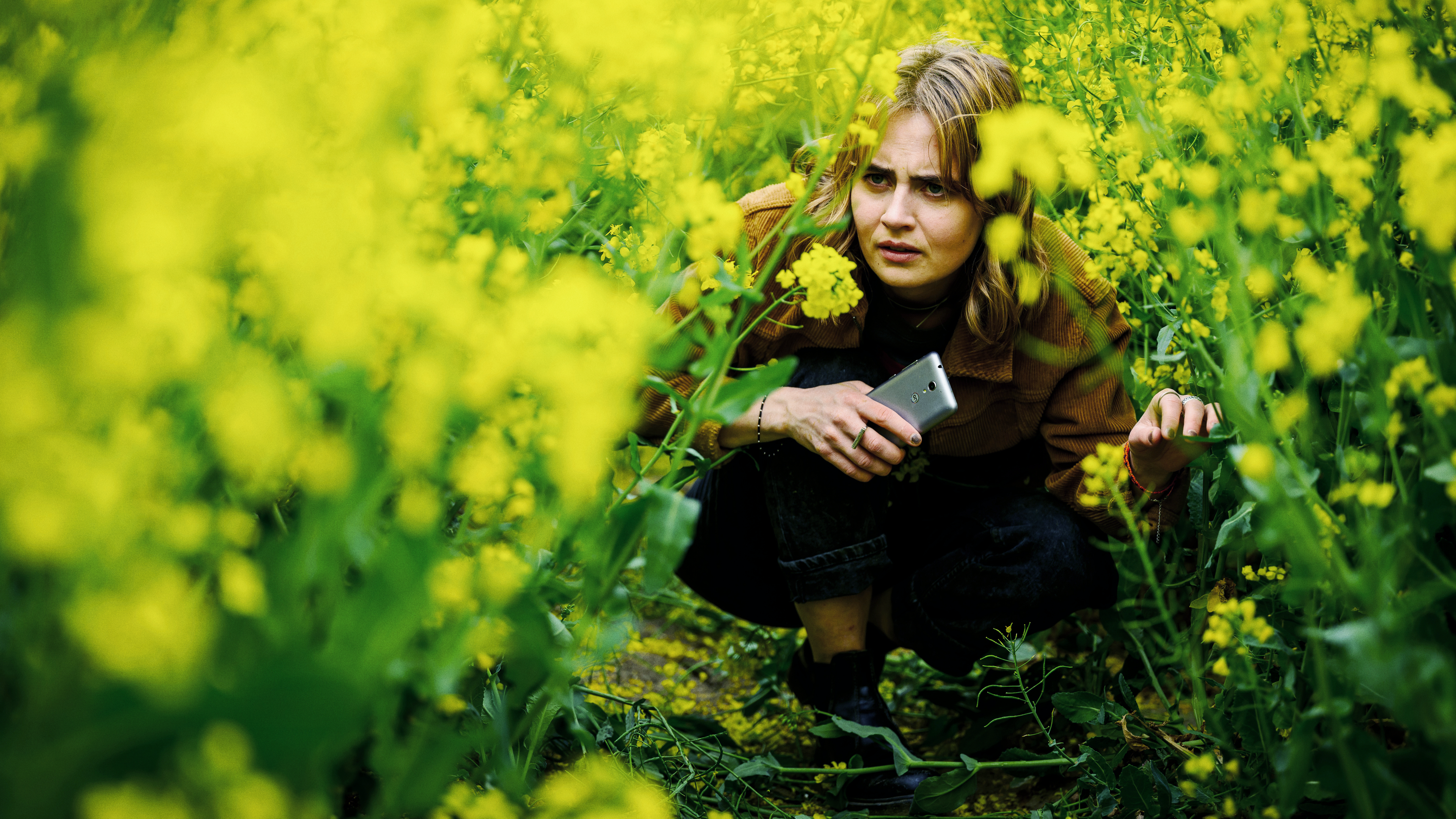 Eine Frau mit einem Handy in der Hand kauert in einem gelb-blühenden Rapsfeld. 