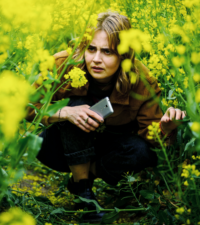 Eine Frau mit einem Handy in der Hand kauert in einem gelb-blühenden Rapsfeld. 
