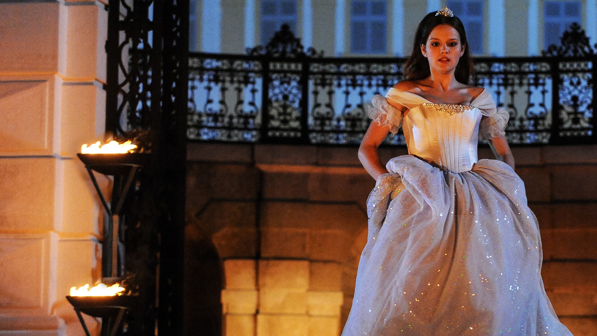 Im Hintergrund das nächtliche Schloss. Aschenputtel (Emilia Schüle), gekleidet in ein schneeweißes Kleid, hat das Kleid in den Hüften etwas angehoben, um den Ball schnell durch die geöffnete, eiserne Toreinfahrt des Schlosses zu verlassen.