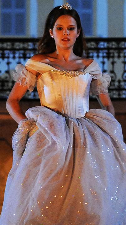 Im Hintergrund das nächtliche Schloss. Aschenputtel (Emilia Schüle), gekleidet in ein schneeweißes Kleid, hat das Kleid in den Hüften etwas angehoben, um den Ball schnell durch die geöffnete, eiserne Toreinfahrt des Schlosses zu verlassen.