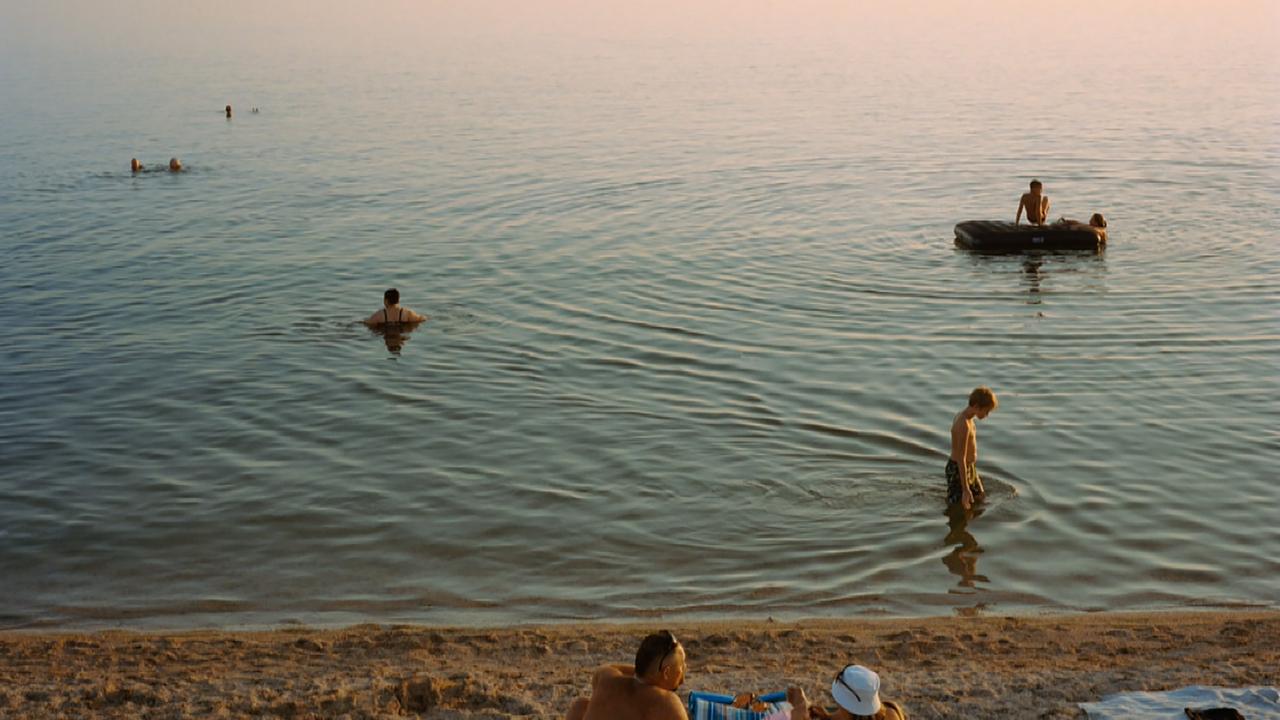 Menschen liegen am Strand in der Sonne, während andere im Meer schwimmen, oder auf Schlachbooten unterwegs sind.