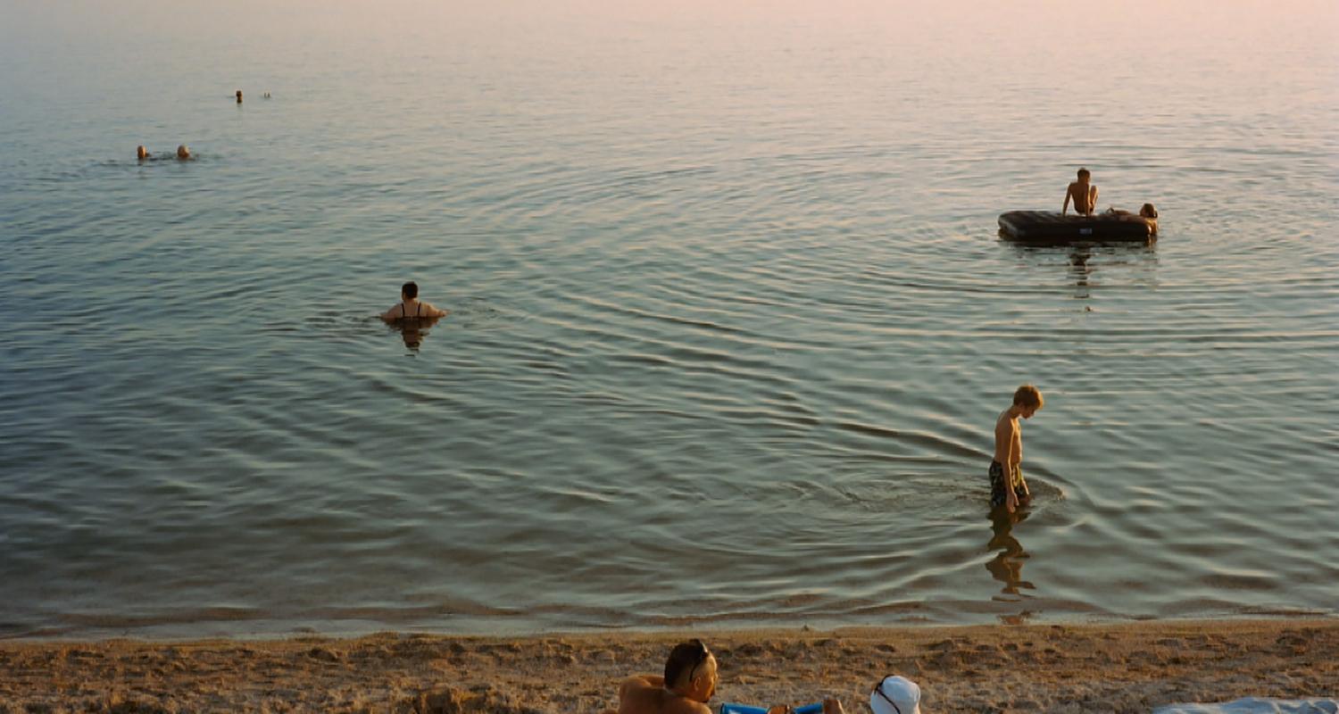 Menschen liegen am Strand in der Sonne, während andere im Meer schwimmen, oder auf Schlachbooten unterwegs sind.