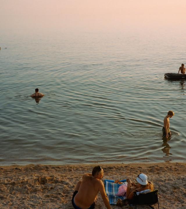 Menschen liegen am Strand in der Sonne, während andere im Meer schwimmen, oder auf Schlachbooten unterwegs sind.