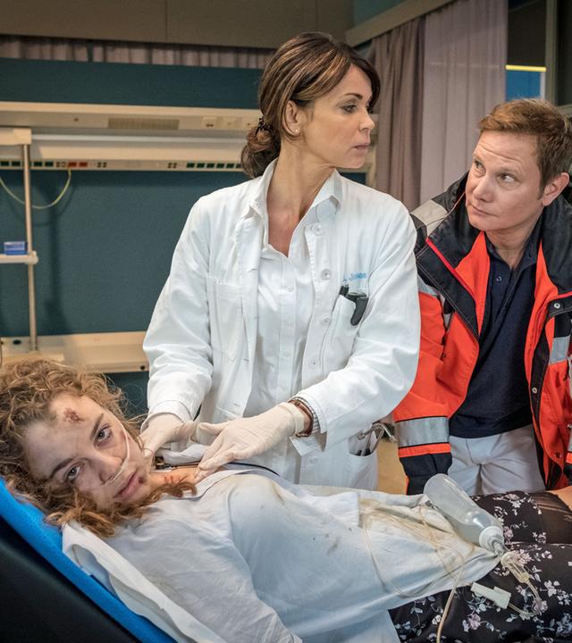 "Notruf Hafenkante - Au-pair": Im Behandlungsraum des EKH stehen Dr. Jonas (Gerit Kling) und Dr. Haase (Fabian Harloff) neben einer Patiententrage, auf der ein junges Mädchen (Amelie Herres) liegt. Das Mädchen hat Blessuren in ihrem Gesicht und ihre Kleidung ist teilweise zerrissen. Es starrt abwesend zur Seite.