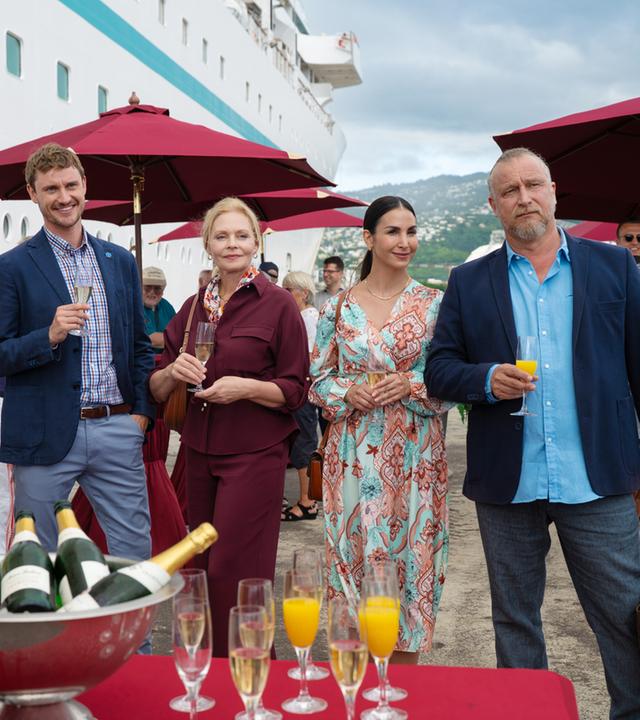  Im Hafen: Esther Sarchow (Susanna Simon), Britta Weigert (Sila Sahin), Victoria Schneider (Luna Baptiste), Erik Milewski (Matthias Komm) und Marco Zurheide (Frédéric Brossier) stehen nebeneinander vor dem Schiff. Sie halten gefüllte Sektgläser in den Händen und wirken glücklich. Vor ihnen steht ein Tisch mit Sektflaschen und -gläsern. Im Hintergrund stehen weitere Passagiere (Komparsen) unter roten Sonnenschirmen an Stehtischen.