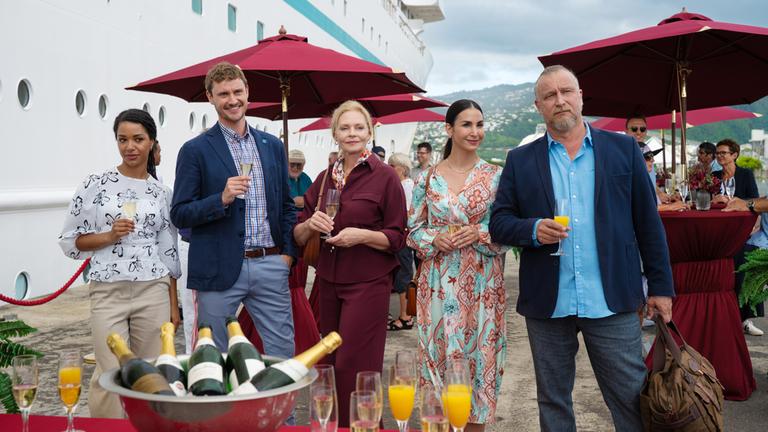  Im Hafen: Esther Sarchow (Susanna Simon), Britta Weigert (Sila Sahin), Victoria Schneider (Luna Baptiste), Erik Milewski (Matthias Komm) und Marco Zurheide (Frédéric Brossier) stehen nebeneinander vor dem Schiff. Sie halten gefüllte Sektgläser in den Händen und wirken glücklich. Vor ihnen steht ein Tisch mit Sektflaschen und -gläsern. Im Hintergrund stehen weitere Passagiere (Komparsen) unter roten Sonnenschirmen an Stehtischen.
