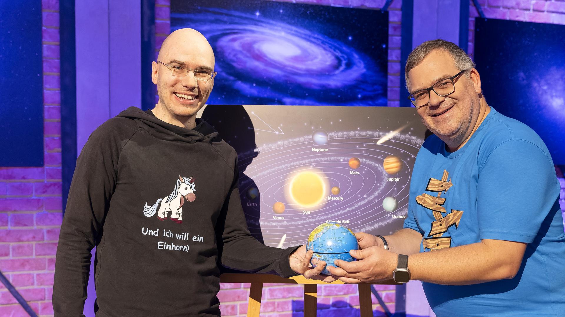 Astronom Mathias Jäger steht neben Elton. Beide halten zusammen lächelnd einen kleinen Globus in den Händen. Hinter ihnen steht eine Tafel mit einer Darstellung des Sonnensystems.