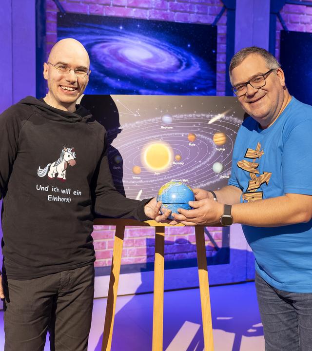 Astronom Mathias Jäger steht neben Elton. Beide halten zusammen lächelnd einen kleinen Globus in den Händen. Hinter ihnen steht eine Tafel mit einer Darstellung des Sonnensystems.