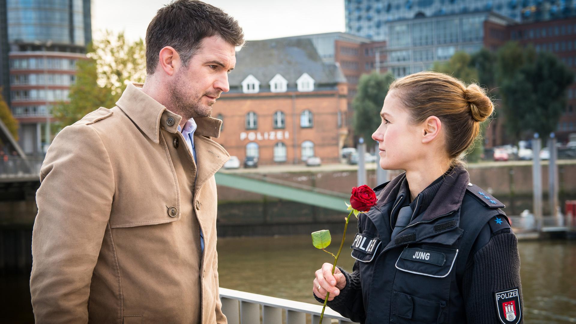 "Notruf Hafenkante - Auf beiden Augen blind": Dr. Philipp Jung (Simon Böer) und seine Frau, die Polizeibeamtin Franzi Jung (Rhea Harder-Vennewald) stehen sich gegenüber und schauen sich an. Franzi hält eine rote Rose in der Hand.