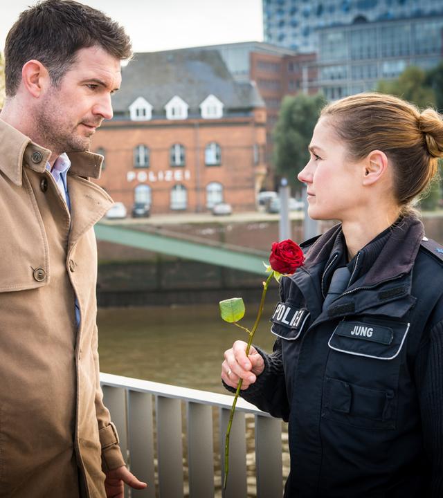 "Notruf Hafenkante - Auf beiden Augen blind": Dr. Philipp Jung (Simon Böer) und seine Frau, die Polizeibeamtin Franzi Jung (Rhea Harder-Vennewald) stehen sich gegenüber und schauen sich an. Franzi hält eine rote Rose in der Hand.