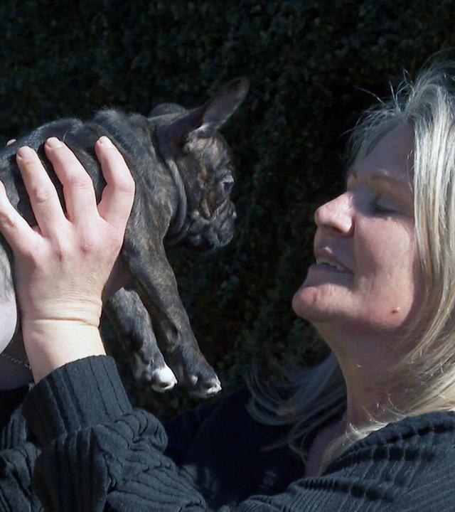 Das Bild zeigt eine Frau, die einen kleinen, dunkel gestromten Hund in ihren Händen hält. Der Hund ist aufrecht, und die Frau schaut ihn liebevoll an, während sie ihn an den Seiten festhält. Die Frau hat lange, blonde Haare und trägt einen schwarzen, langärmligen Pullover. Im Hintergrund sind grüne Sträucher zu sehen, die eine natürliche Umgebung schaffen. Das Licht der Szene wirkt hell und freundlich, was eine positive Atmosphäre vermittelt.