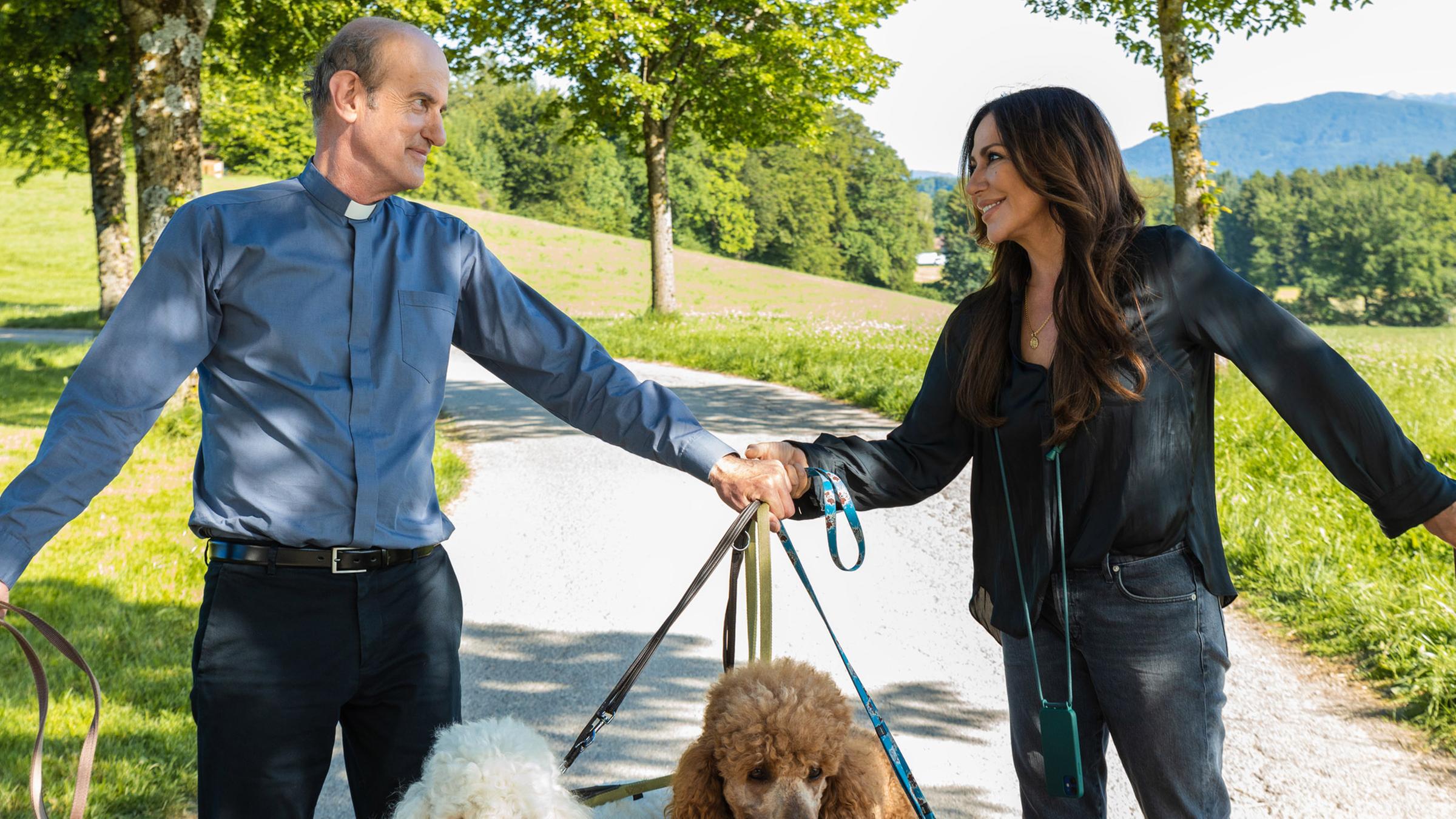 Katja Baumann (Simone Thomalla) und Pfarrer Sonnleitner (Johannes Herrschmann) gehen mit mehreren Hunden an der Leine zwischen Wiesen spazieren.