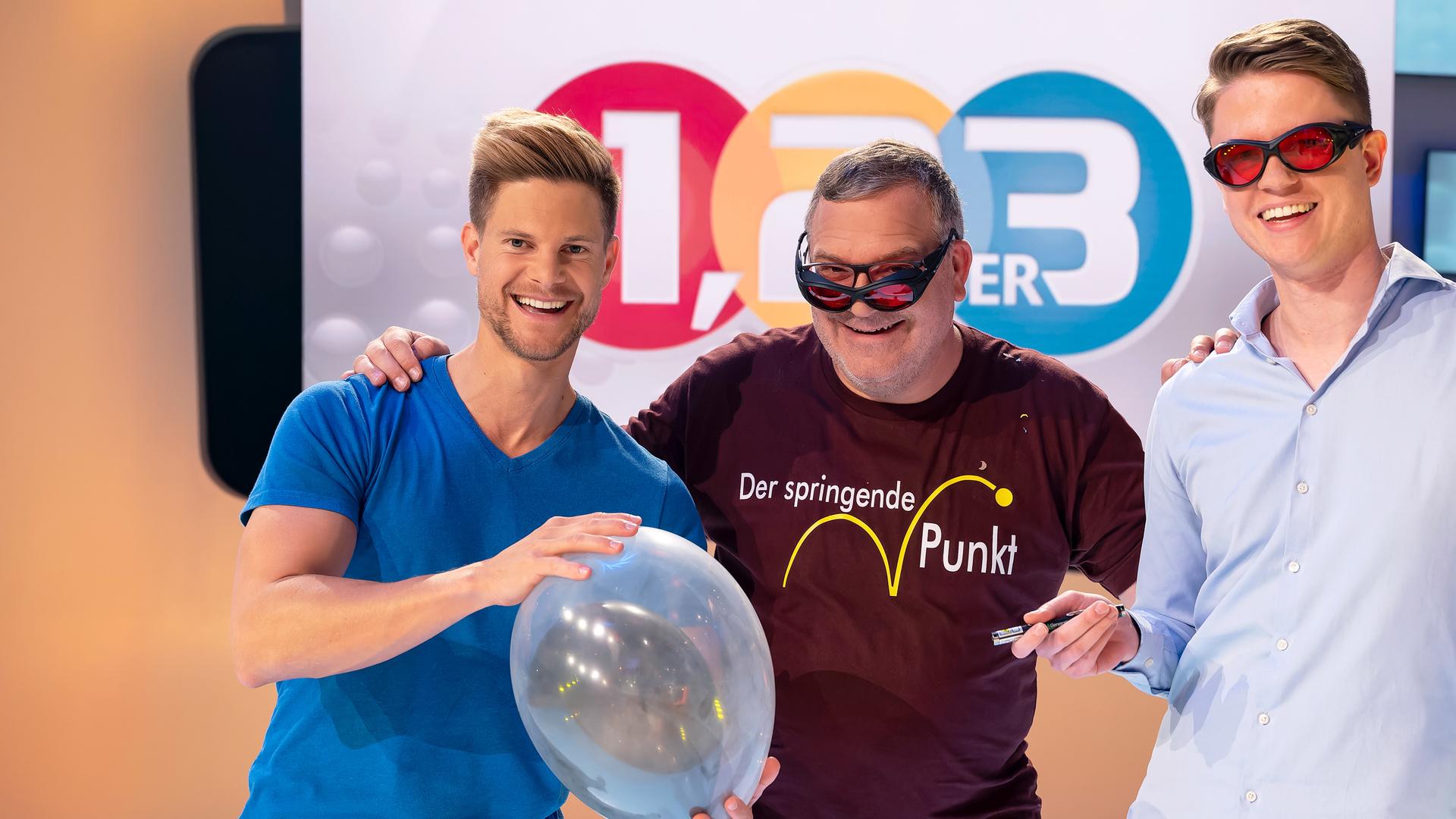 Elton steht in der Mitte zwischen Dr. Philip Häusser und Moritz Heindl. Dr. Philip Häusser hält einen durchsichtigen Luftballon in den Händen, in dem sich ein weiterer, schwarzer Luftballon befindet. Moritz Heindl hält einen Laserpointer in der rechten Hand. Er und Elton tragen jeweils eine schwarze Schutzbrille mit roten Gläsern.