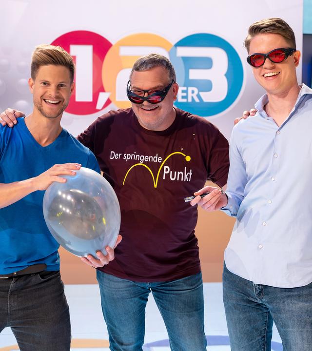 Elton steht in der Mitte zwischen Dr. Philip Häusser und Moritz Heindl. Dr. Philip Häusser hält einen durchsichtigen Luftballon in den Händen, in dem sich ein weiterer, schwarzer Luftballon befindet. Moritz Heindl hält einen Laserpointer in der rechten Hand. Er und Elton tragen jeweils eine schwarze Schutzbrille mit roten Gläsern.