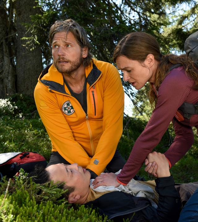 "Die Bergretter: Auf der Jagd": Auf einem begrünten Waldboden liegt Tom (Johannes Ahn) mit bleichem Gesicht. Er blickt zu Nina (Josephin Busch) auf, die neben ihm kniet, seine Hand hält und ein Tuch auf seine Brust drückt. Ihr gegenüber kniet Markus (Sebastian Ströbel), der angespannt in den Himmel schaut.
