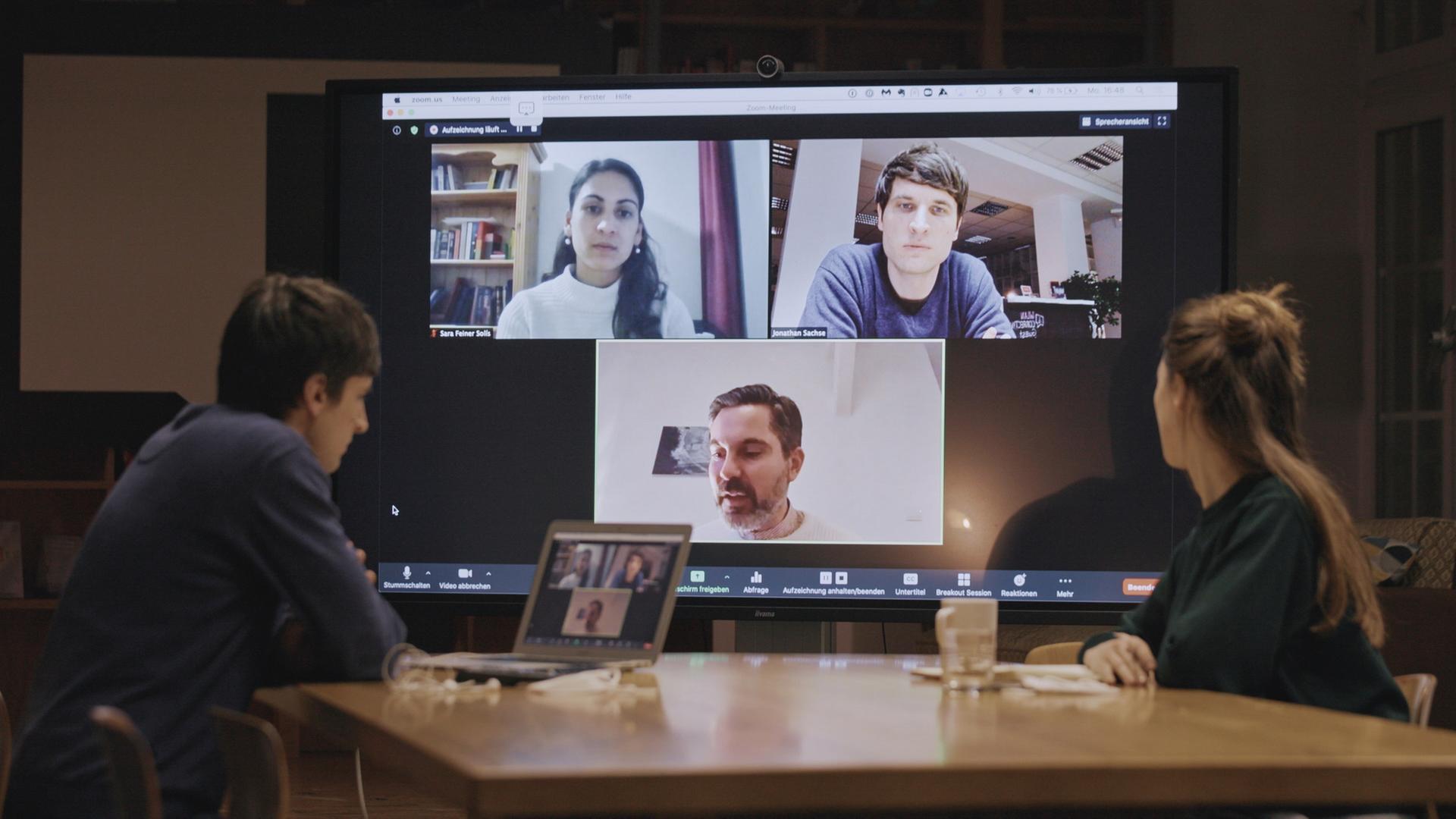 Eine Frau und ein Mann sitzen an einem Konferenztisch mit einem großen Monitor im Hintergrund, auf den sie schauen und auf dem weitere drei Personen, eine Frau und zwei Männer, in einer Videokonferenz zu sehen sind.