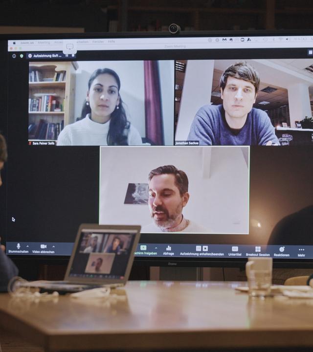Eine Frau und ein Mann sitzen an einem Konferenztisch mit einem großen Monitor im Hintergrund, auf den sie schauen und auf dem weitere drei Personen, eine Frau und zwei Männer, in einer Videokonferenz zu sehen sind.