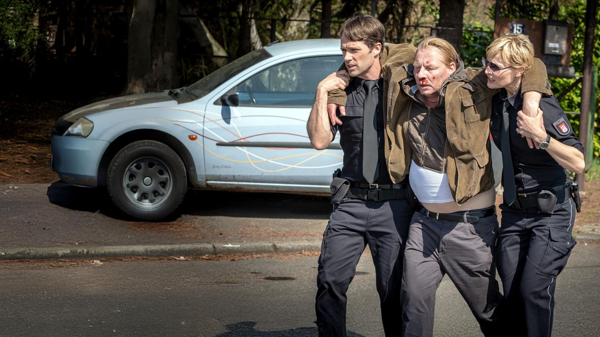 "Notruf Hafenkante - Auf der Straße": Die Polizisten Mattes Seeler (Matthias Schloo) und Melanie Hansen (Sanna Englund) stützen Heinrich Frank (Ben Becker), der zwischen ihnen geht. Heinrich Frank hat jeweils einen Arm über die Schulter eines Polizisten gelegt. Er hat Verletzungen im Gesicht, die bluten.
