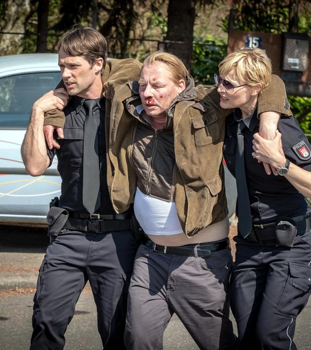 "Notruf Hafenkante - Auf der Straße": Die Polizisten Mattes Seeler (Matthias Schloo) und Melanie Hansen (Sanna Englund) stützen Heinrich Frank (Ben Becker), der zwischen ihnen geht. Heinrich Frank hat jeweils einen Arm über die Schulter eines Polizisten gelegt. Er hat Verletzungen im Gesicht, die bluten.