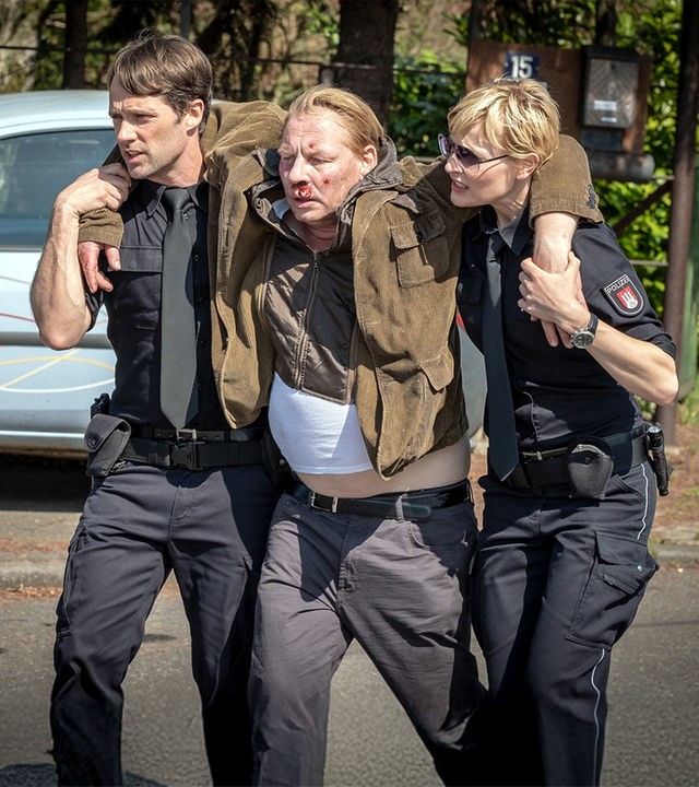 "Notruf Hafenkante - Auf der Straße": Die Polizisten Mattes Seeler (Matthias Schloo) und Melanie Hansen (Sanna Englund) stützen Heinrich Frank (Ben Becker), der zwischen ihnen geht. Heinrich Frank hat jeweils einen Arm über die Schulter eines Polizisten gelegt. Er hat Verletzungen im Gesicht, die bluten.