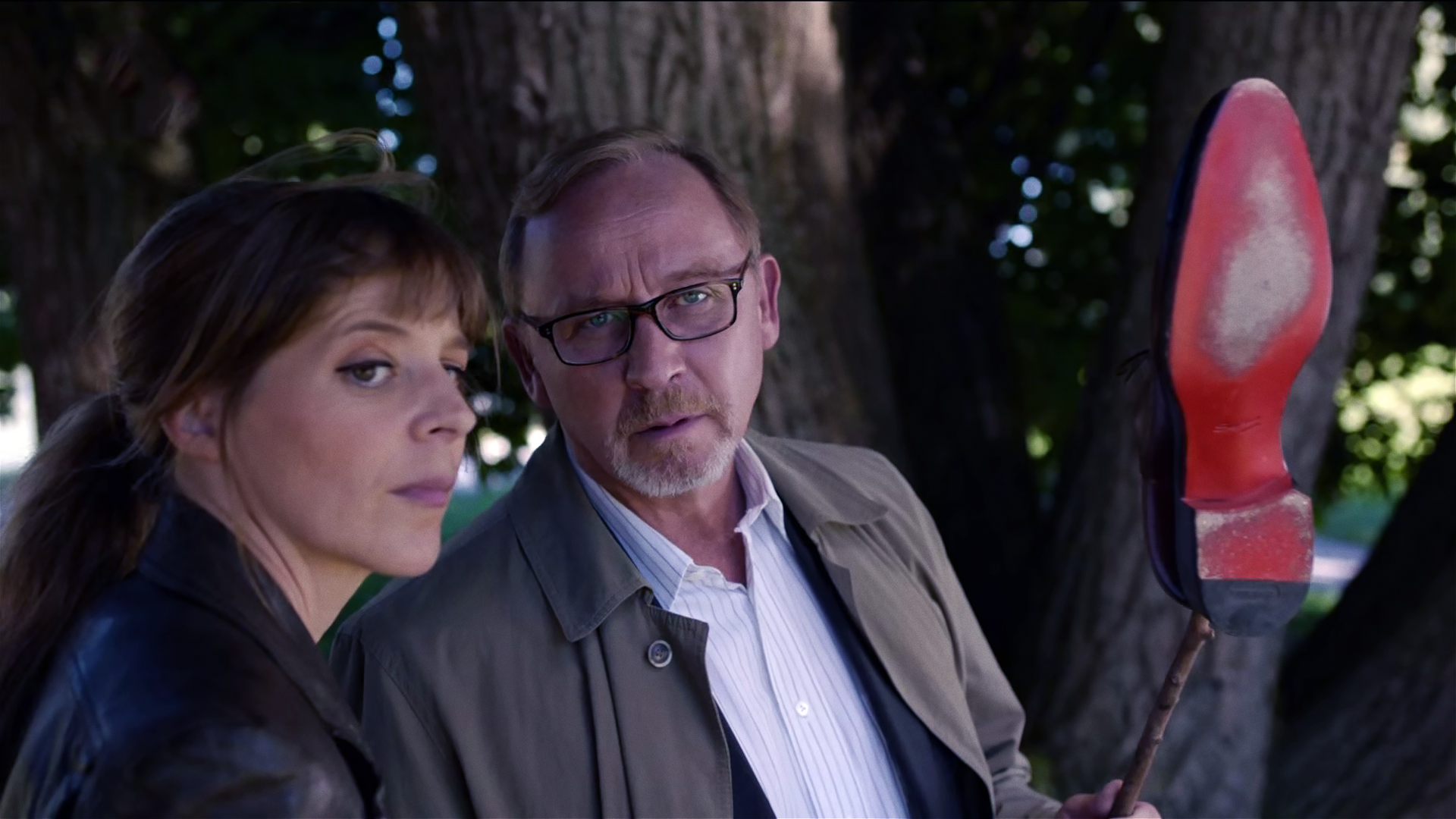 Angelika Flierl (Bernadette Heerwagen) und Ludwig Schaller (Alexander Held) betrachten einen Herrenschuh mit roter Sohle, den Schaller auf einem Stock aufgehoben hat.