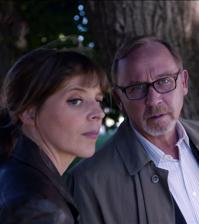 Angelika Flierl (Bernadette Heerwagen) und Ludwig Schaller (Alexander Held) betrachten einen Herrenschuh mit roter Sohle, den Schaller auf einem Stock aufgehoben hat.