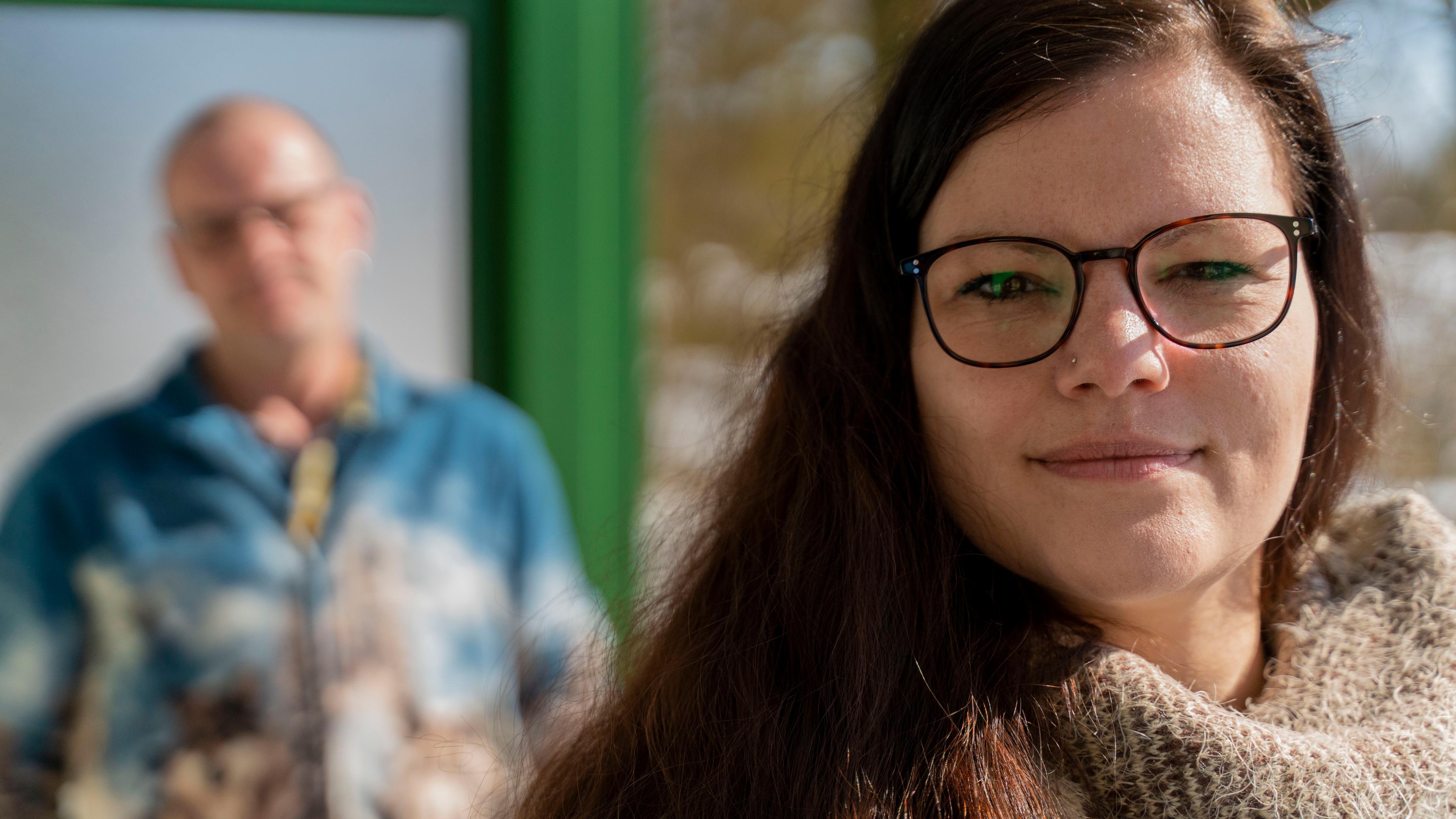 Claudia schaut in die Kamera. Im Hintergrund ist unscharf ihr Vater zu sehen.