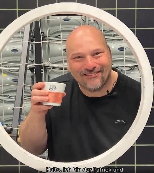 Screenshot ZDF-Aufnahmeleiter Patrick mit erhobener Mainzelmännchen-Tasse blickt von hinten durch Torwandloch und lächelt.