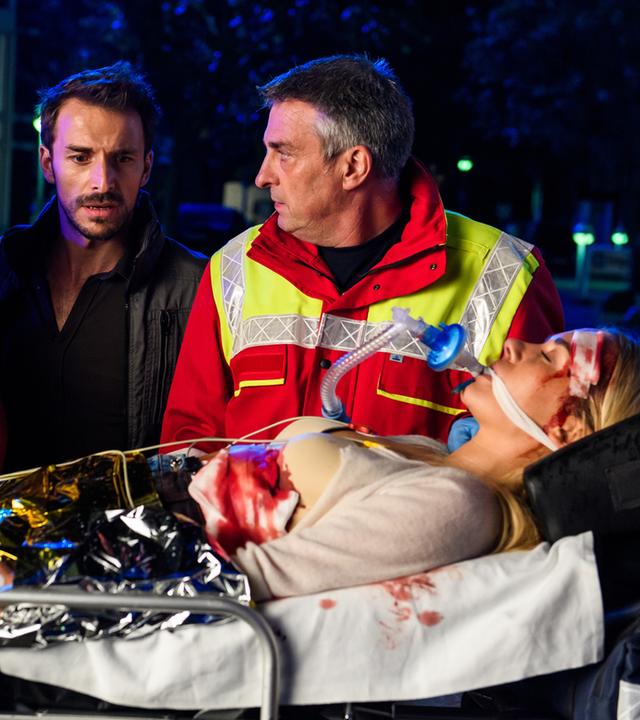 "Bettys Diagnose - Auf Messers Schneide": Hanna Winter (Marie Zielcke) und Dr. Frank Stern (Max Alberti) schauen entsetzt auf die verletzte Betty (Annina Hellenthal), die gerade von einem Notarzt (Komparse) in die Karlsklinik eingeliefert wird.