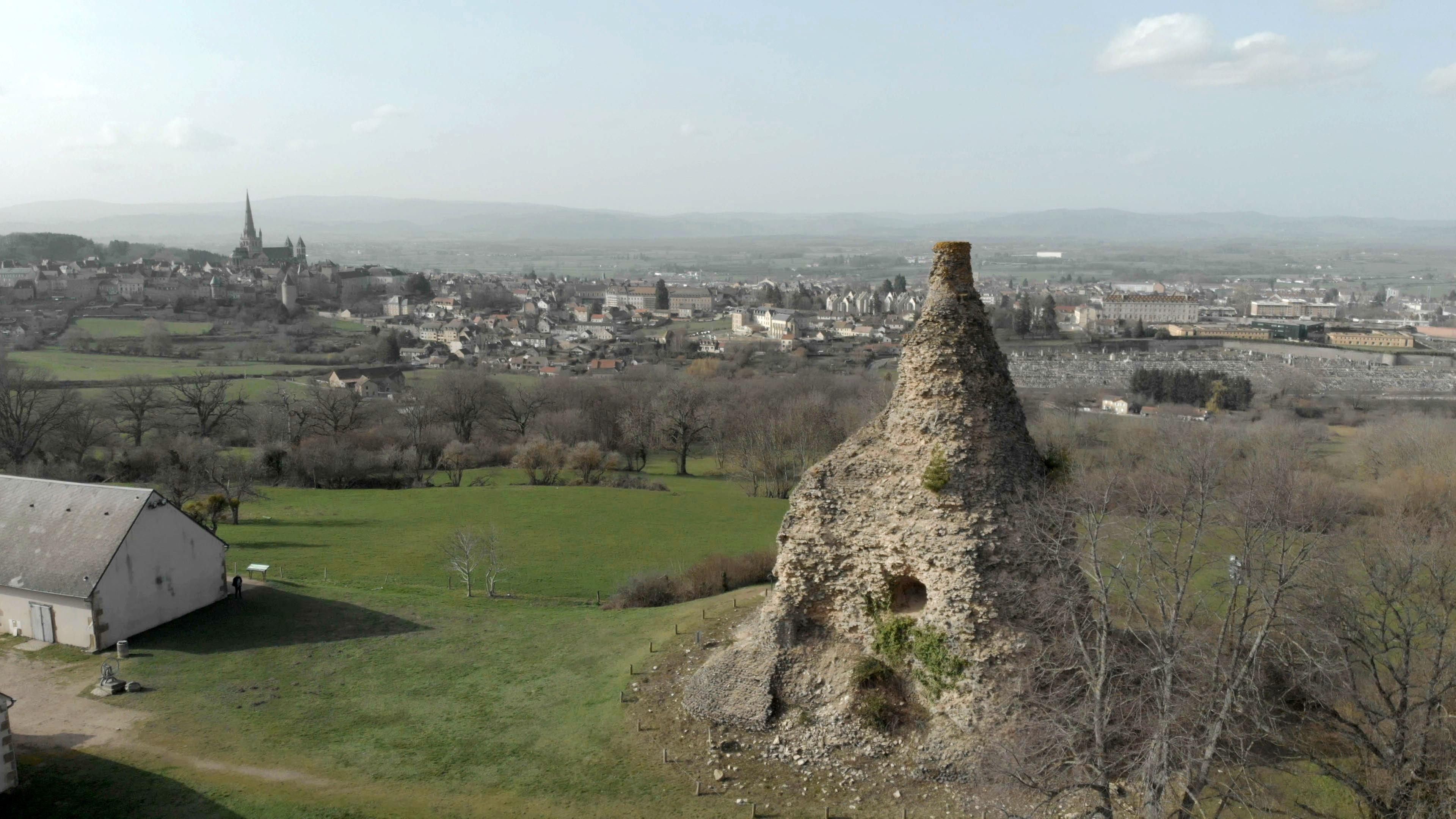 Im Vordergrund ein pyramidenförmiges halb verfallenes Bauwerk. Im Hintergrund eine Kleinstadt und sanfte Hügel.