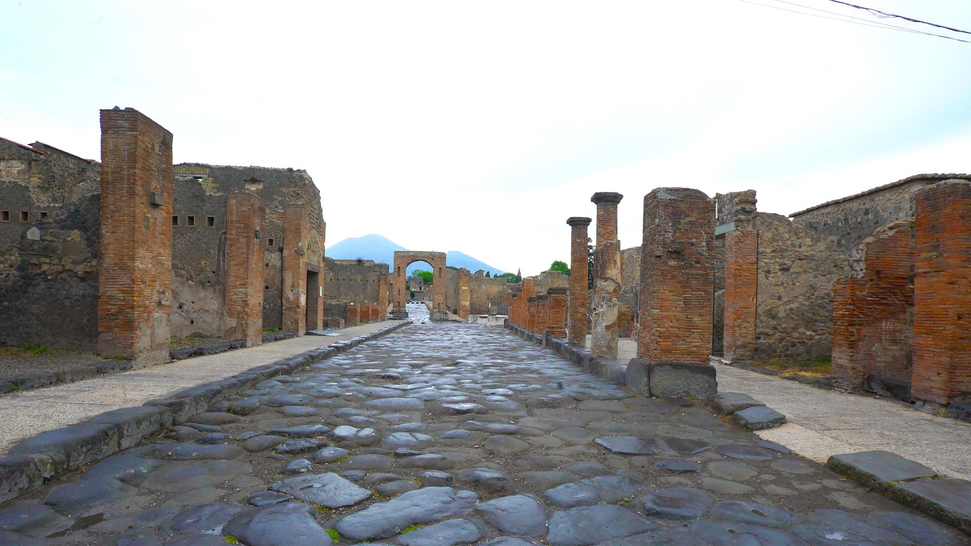  Eine gepflasterte Straße von Pompeji. Am Straßenrand sind Ruinen von Häusern zu sehen.
