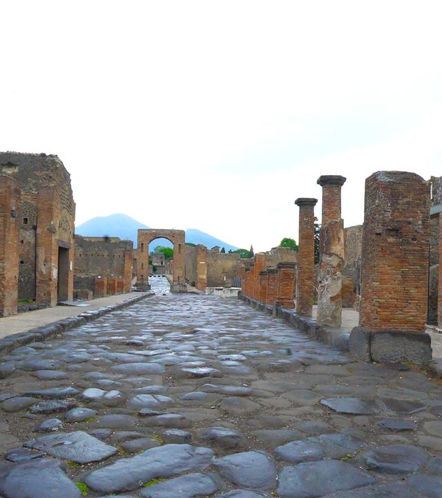  Eine gepflasterte Straße von Pompeji. Am Straßenrand sind Ruinen von Häusern zu sehen.