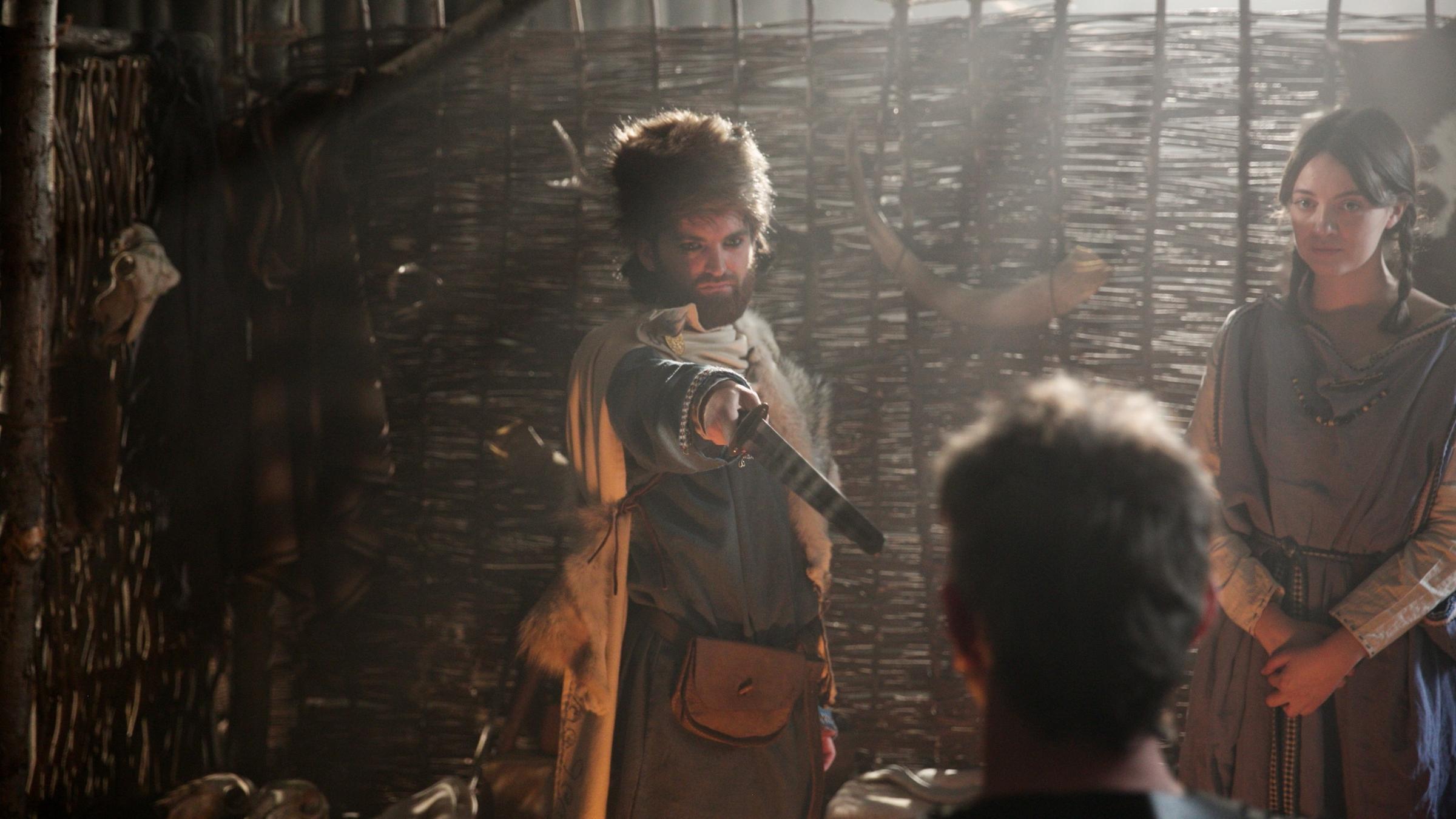 Ein Mann mit Tierfell auf dem Kopf zeigt mit einem Schwert auf einen anderen Mann. Neben ihnen steht ein Mädchen mit Zöpfen.