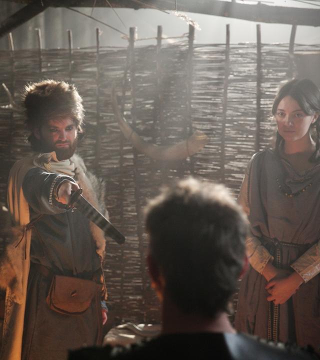 Ein Mann mit Tierfell auf dem Kopf zeigt mit einem Schwert auf einen anderen Mann. Neben ihnen steht ein Mädchen mit Zöpfen.