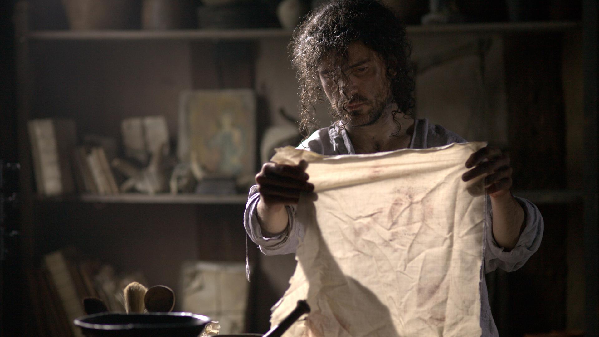 "Aufgedeckt - Rätsel der Geschichte: Das Grabtuch von Turin": Nachgestellte Szene: Ein Mann sitzt in einem Atelier und hält einen Fetzen eines Leinentuchs in den Händen.