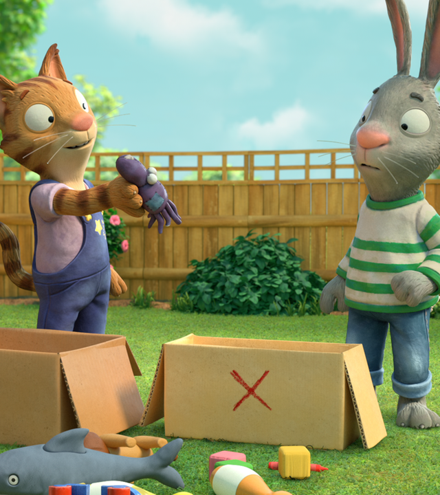 Frankie zeigt Pip im Garten ein altes Spielzeug. Pip überlegt, ob er es behalten oder wegwerfen möchte. Vor ihnen steht jeweils ein Pappkarton zum Ausmisten.