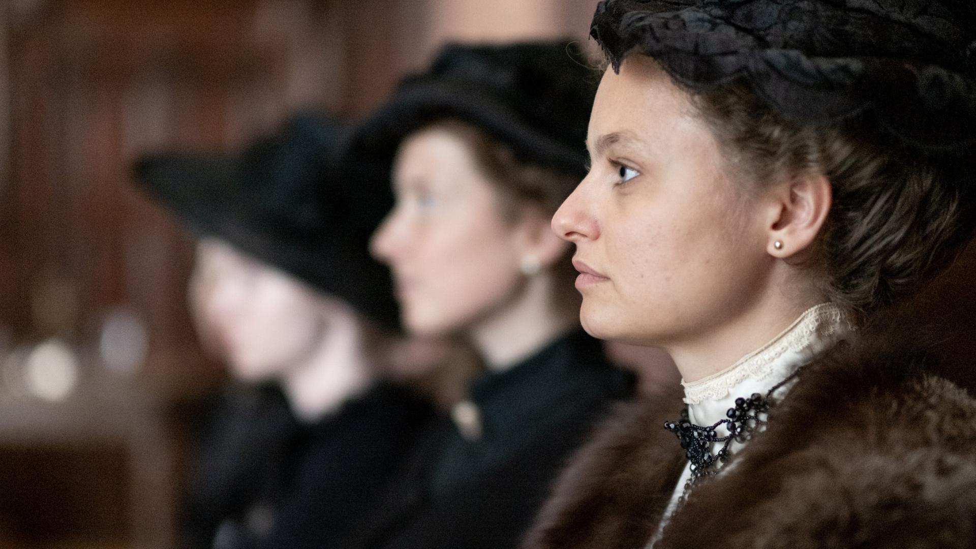 Das Bild zeigt eine Szene aus einem historischen Films. Im Vordergrund sieht man eine Frau in einem eleganten, historischen Kostüm, die zur Seite schaut. Sie trägt einen dunklen Hut und hat ihre Haare hochgesteckt. Ihr Outfit ist verziert mit einer auffälligen Halskette. Hinter ihr sind zwei weitere Frauen sichtbar, die ebenfalls ähnliche historische Kleidung tragen, jedoch nur teilweise erkennbar sind, da ihre Gesichter zum Teil im Schatten liegen.  Die Umgebung scheint ein geräumiger Innenraum zu sein, möglicherweise ein Gerichtssaal oder ein ähnlicher Ort, der mit dunklem Holz verkleidet ist. Die Stimmung des Bildes kann als ernst und nachdenklich beschrieben werden, passend zum Kontext des Films, der sich mit Themen wie Frauenhandel und gesellschaftlichen Missständen zu Beginn des 20. Jahrhunderts beschäftigt.