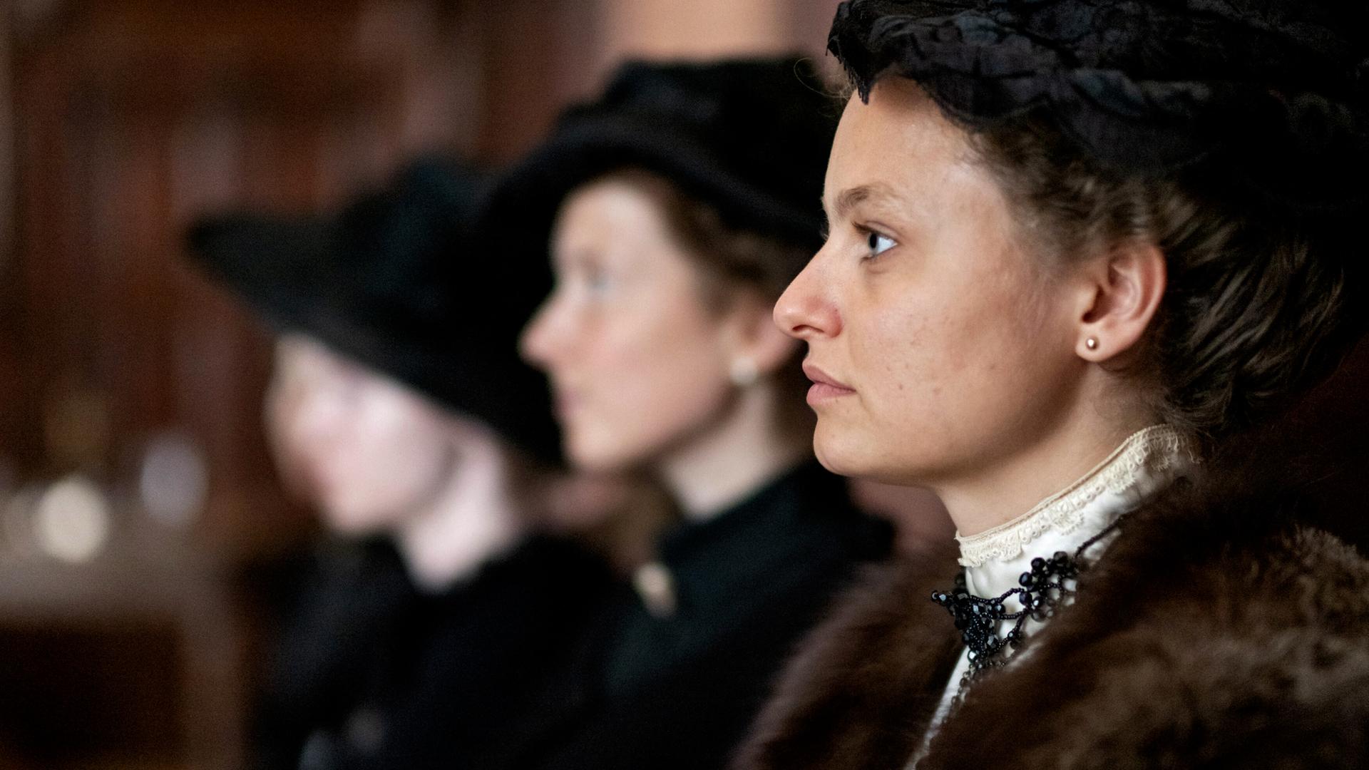 Das Bild zeigt eine Szene aus einem historischen Films. Im Vordergrund sieht man eine Frau in einem eleganten, historischen Kostüm, die zur Seite schaut. Sie trägt einen dunklen Hut und hat ihre Haare hochgesteckt. Ihr Outfit ist verziert mit einer auffälligen Halskette. Hinter ihr sind zwei weitere Frauen sichtbar, die ebenfalls ähnliche historische Kleidung tragen, jedoch nur teilweise erkennbar sind, da ihre Gesichter zum Teil im Schatten liegen.  Die Umgebung scheint ein geräumiger Innenraum zu sein, möglicherweise ein Gerichtssaal oder ein ähnlicher Ort, der mit dunklem Holz verkleidet ist. Die Stimmung des Bildes kann als ernst und nachdenklich beschrieben werden, passend zum Kontext des Films, der sich mit Themen wie Frauenhandel und gesellschaftlichen Missständen zu Beginn des 20. Jahrhunderts beschäftigt.