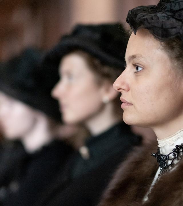 Das Bild zeigt eine Szene aus einem historischen Films. Im Vordergrund sieht man eine Frau in einem eleganten, historischen Kostüm, die zur Seite schaut. Sie trägt einen dunklen Hut und hat ihre Haare hochgesteckt. Ihr Outfit ist verziert mit einer auffälligen Halskette. Hinter ihr sind zwei weitere Frauen sichtbar, die ebenfalls ähnliche historische Kleidung tragen, jedoch nur teilweise erkennbar sind, da ihre Gesichter zum Teil im Schatten liegen.  Die Umgebung scheint ein geräumiger Innenraum zu sein, möglicherweise ein Gerichtssaal oder ein ähnlicher Ort, der mit dunklem Holz verkleidet ist. Die Stimmung des Bildes kann als ernst und nachdenklich beschrieben werden, passend zum Kontext des Films, der sich mit Themen wie Frauenhandel und gesellschaftlichen Missständen zu Beginn des 20. Jahrhunderts beschäftigt.