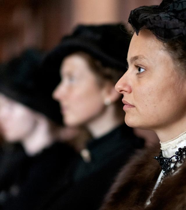 Das Bild zeigt eine Szene aus einem historischen Films. Im Vordergrund sieht man eine Frau in einem eleganten, historischen Kostüm, die zur Seite schaut. Sie trägt einen dunklen Hut und hat ihre Haare hochgesteckt. Ihr Outfit ist verziert mit einer auffälligen Halskette. Hinter ihr sind zwei weitere Frauen sichtbar, die ebenfalls ähnliche historische Kleidung tragen, jedoch nur teilweise erkennbar sind, da ihre Gesichter zum Teil im Schatten liegen.  Die Umgebung scheint ein geräumiger Innenraum zu sein, möglicherweise ein Gerichtssaal oder ein ähnlicher Ort, der mit dunklem Holz verkleidet ist. Die Stimmung des Bildes kann als ernst und nachdenklich beschrieben werden, passend zum Kontext des Films, der sich mit Themen wie Frauenhandel und gesellschaftlichen Missständen zu Beginn des 20. Jahrhunderts beschäftigt.