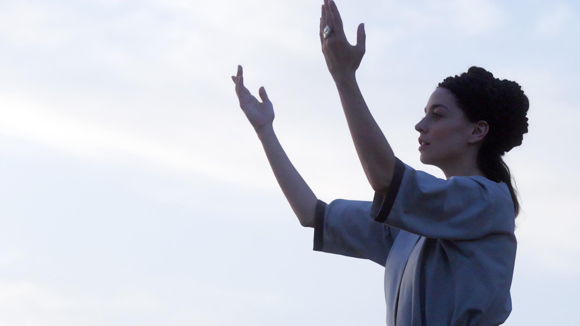 Spielszene: Sijama (Rania Athanasopoulou) steht am Heiligen Berg und spricht ein Gebet. Sie hat ihre Hände gen Himmel erhoben.