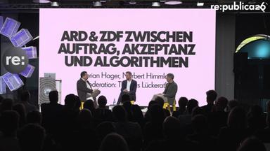 Auf einer Bühne der re:publica 26 sitzen Moderator Thomas Lückerath sowie die Intendanten Florian Hager (HR) und Norbert Himmler (ZDF) in einer Diskussionsrunde vor Publikum. Hinter ihnen ist groß der Titel „ARD & ZDF zwischen Auftrag, Akzeptanz und Algorithmen“ zu sehen.