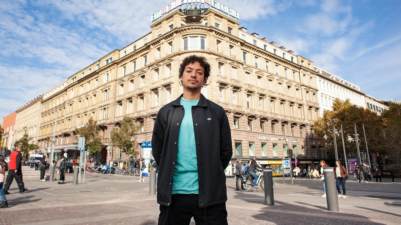 Ein junger Mann steht in einer Großstadt vor einem großen Gebäude an einer Kreuzung.