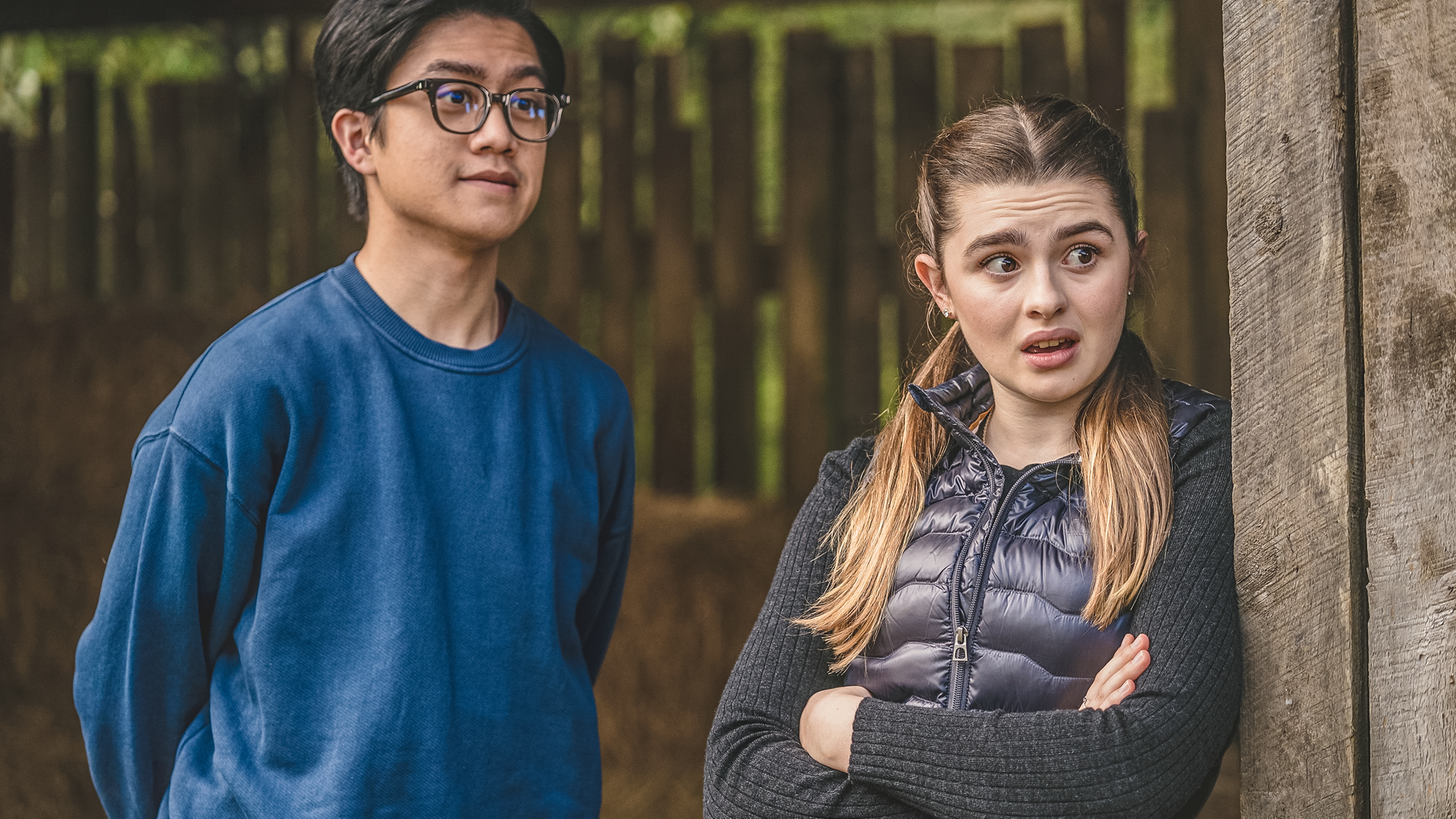 Draußen lehnt Natascha mit verschränkten Armen an einem Holzpfeiler. Sie schaut überrascht mit offenem Mund und großen Augen nach rechts. Neben ihr steht Caleb. Er hat die Armen auf dem Rücken und schaut in dieselbe Richtung.