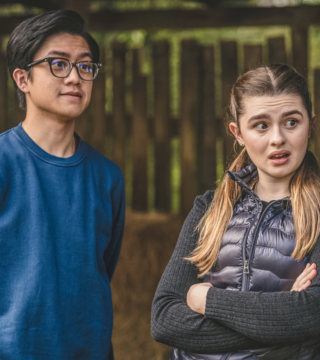 Draußen lehnt Natascha mit verschränkten Armen an einem Holzpfeiler. Sie schaut überrascht mit offenem Mund und großen Augen nach rechts. Neben ihr steht Caleb. Er hat die Armen auf dem Rücken und schaut in dieselbe Richtung.