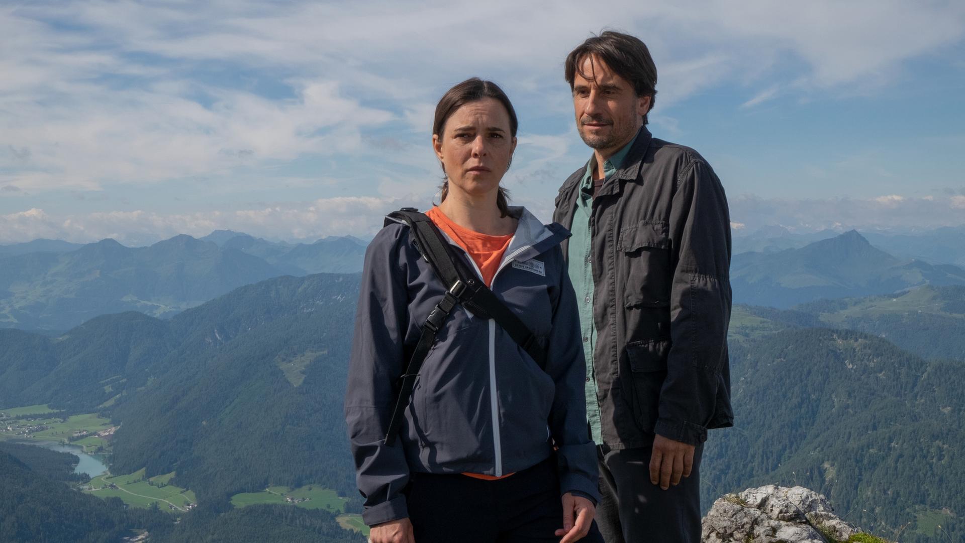 Ein Mann und eine Frau stehen erhöht auf einem Berg und schauen an der Kamera vorbei angespannt in die Ferne. Hinter ihnen Bergpanorama.