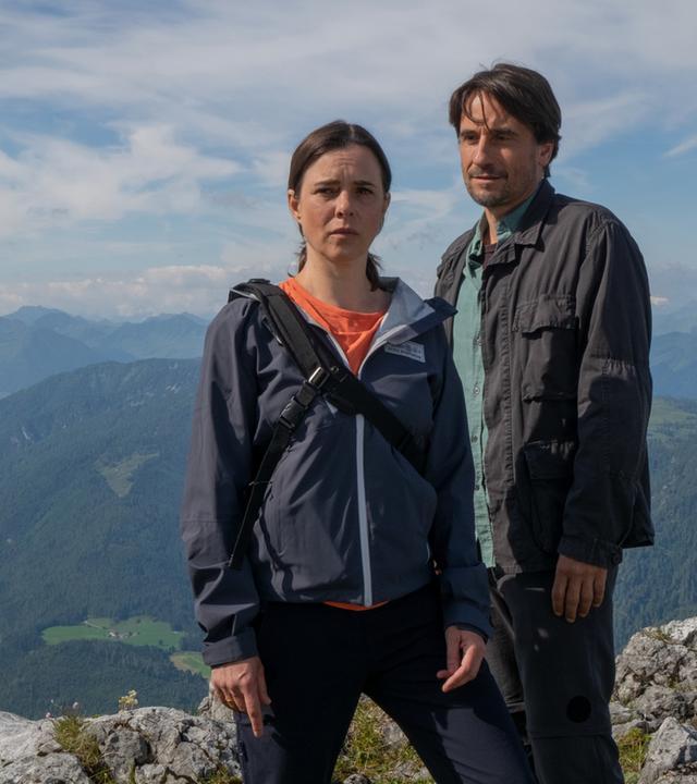 Dr. Martin Gruber (Hans Sigl) und Tim Hauser (Oliver Mommsen) stehen erhöht auf einem Berg und schauen an der Kamera vorbei angespannt in die Ferne. Hinter ihnen Bergpanorama.