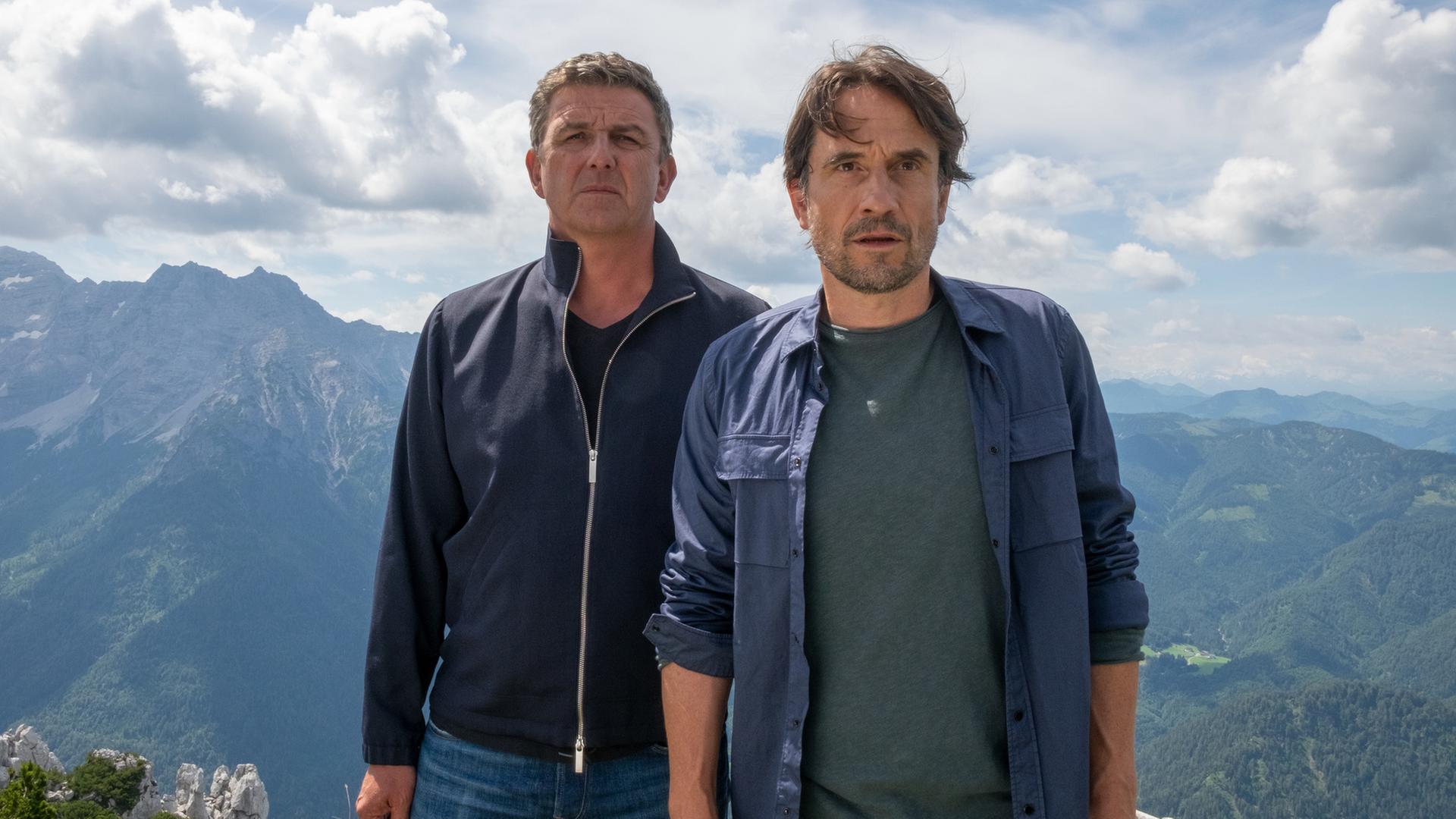 "Der Bergdoktor: Aus Mut gemacht": Dr. Martin Gruber (Hans Sigl) und Tim Hauser (Oliver Mommsen) stehen erhöht auf einem Berg und schauen an der Kamera vorbei angespannt in die Ferne. Hinter ihnen Bergpanorama.